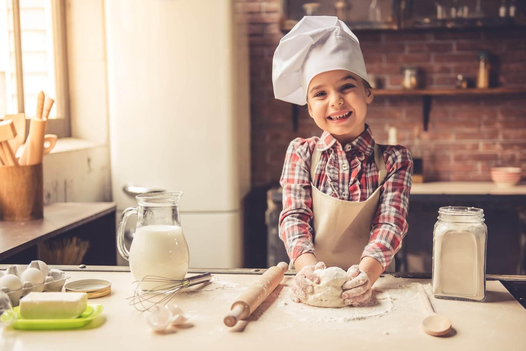 Child in chef hat smiling, with dough and baking utensils