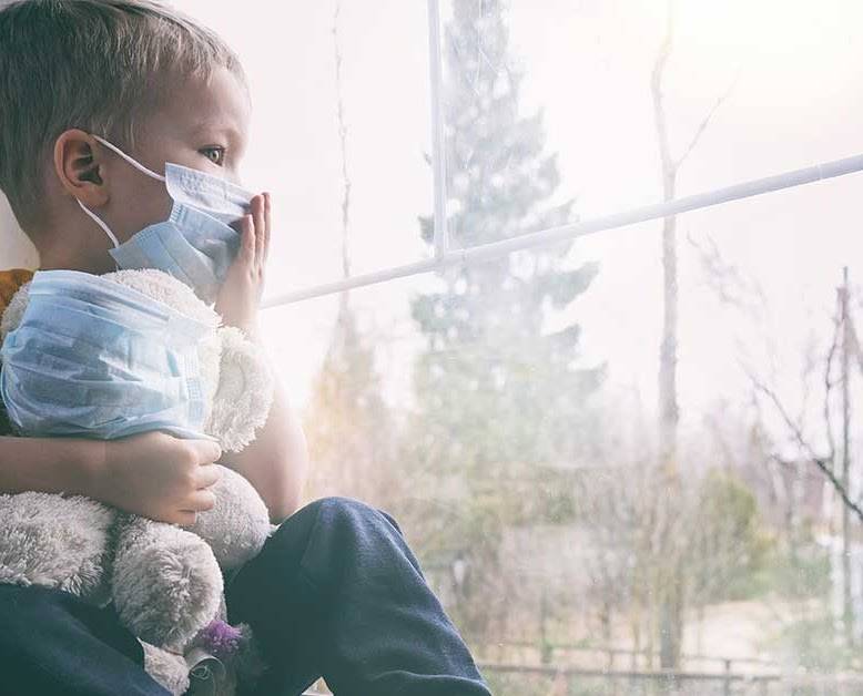 Child with mask holding bear with mask near window, looking outside at sun