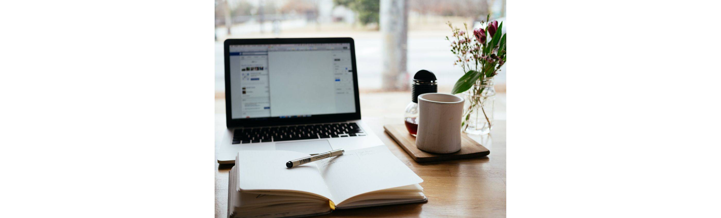 Open notebook in front of a laptop doing research paper next to cup of tea and dried flowers