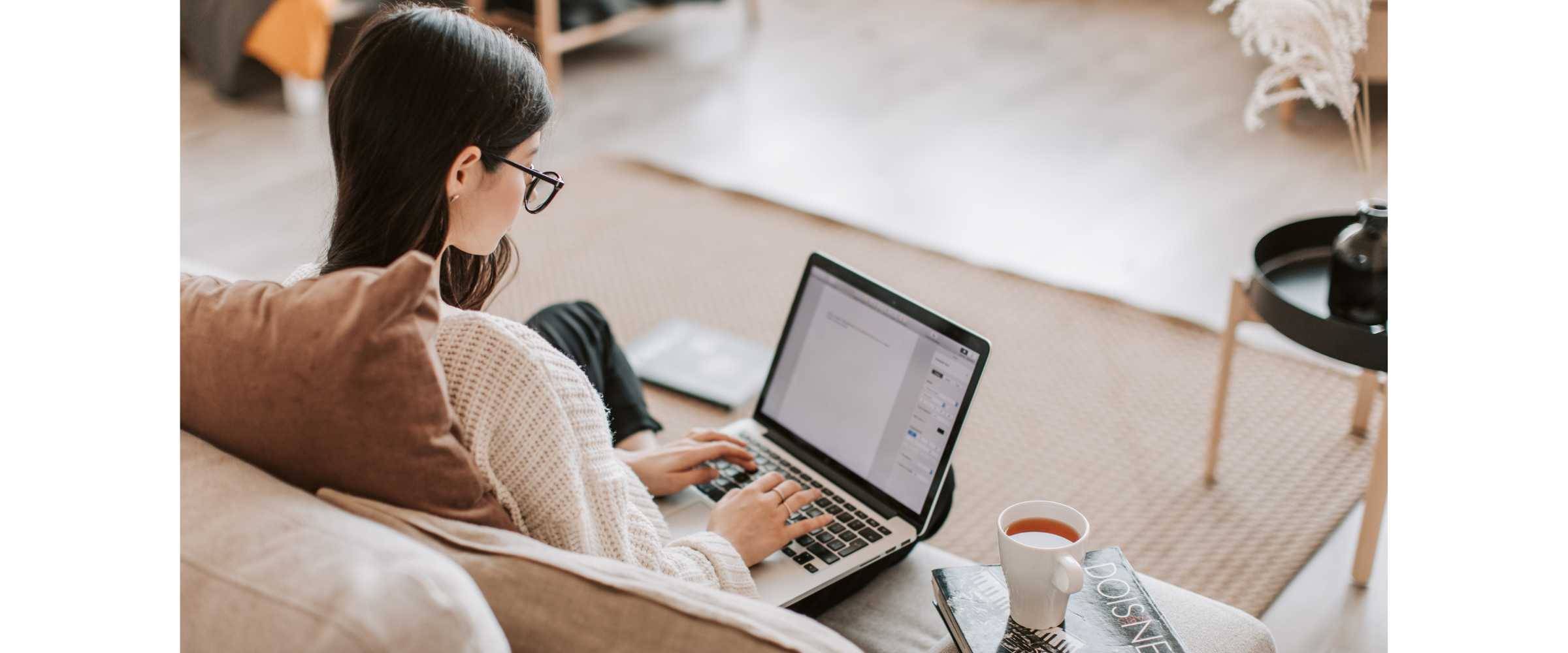 young woman with dark hair and glasses typing on a Macbook next to a cup of tea and a book